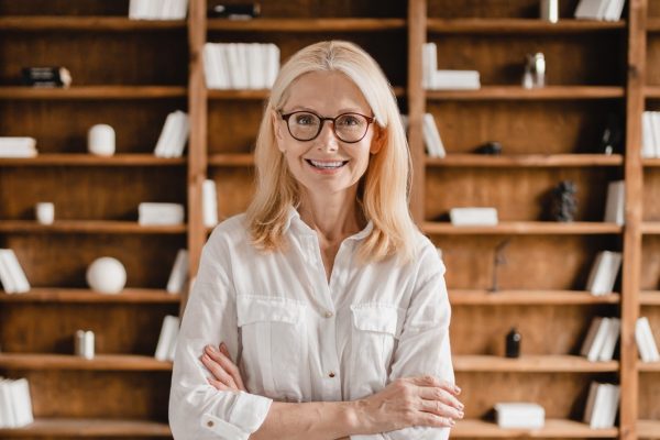Smart caucasian middle-aged mature businesswoman librarian psychologist boss teacher in glasses with arms crossed looking at the camera in library office