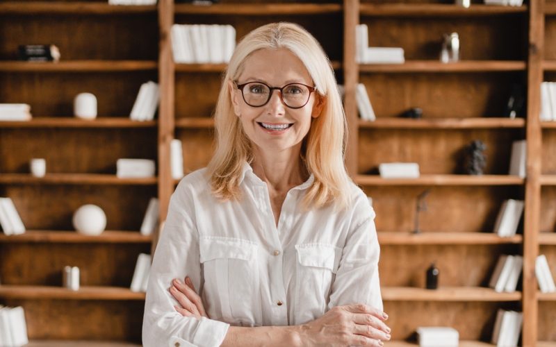 Smart caucasian middle-aged mature businesswoman librarian psychologist boss teacher in glasses with arms crossed looking at the camera in library office